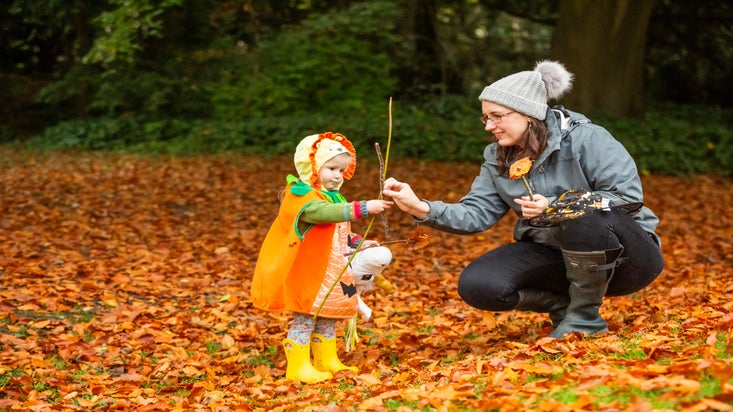 Adult with child dressed in Halloween costume, crouching down surrounded by autumn leaves